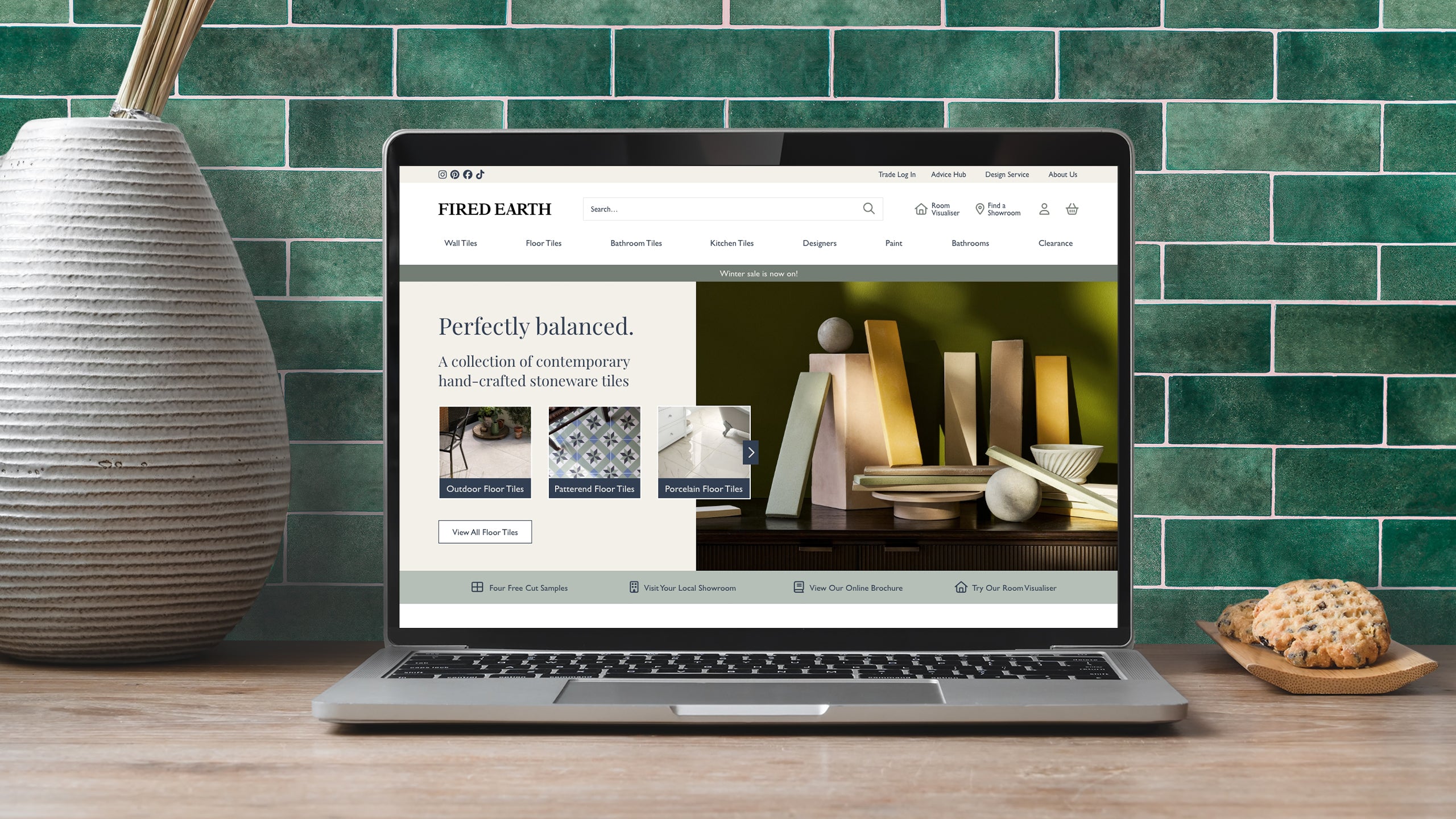 Laptop displaying a Shopify Plus website for a tiles company, Fired Earth, on a wooden table with a green tiled wall background.