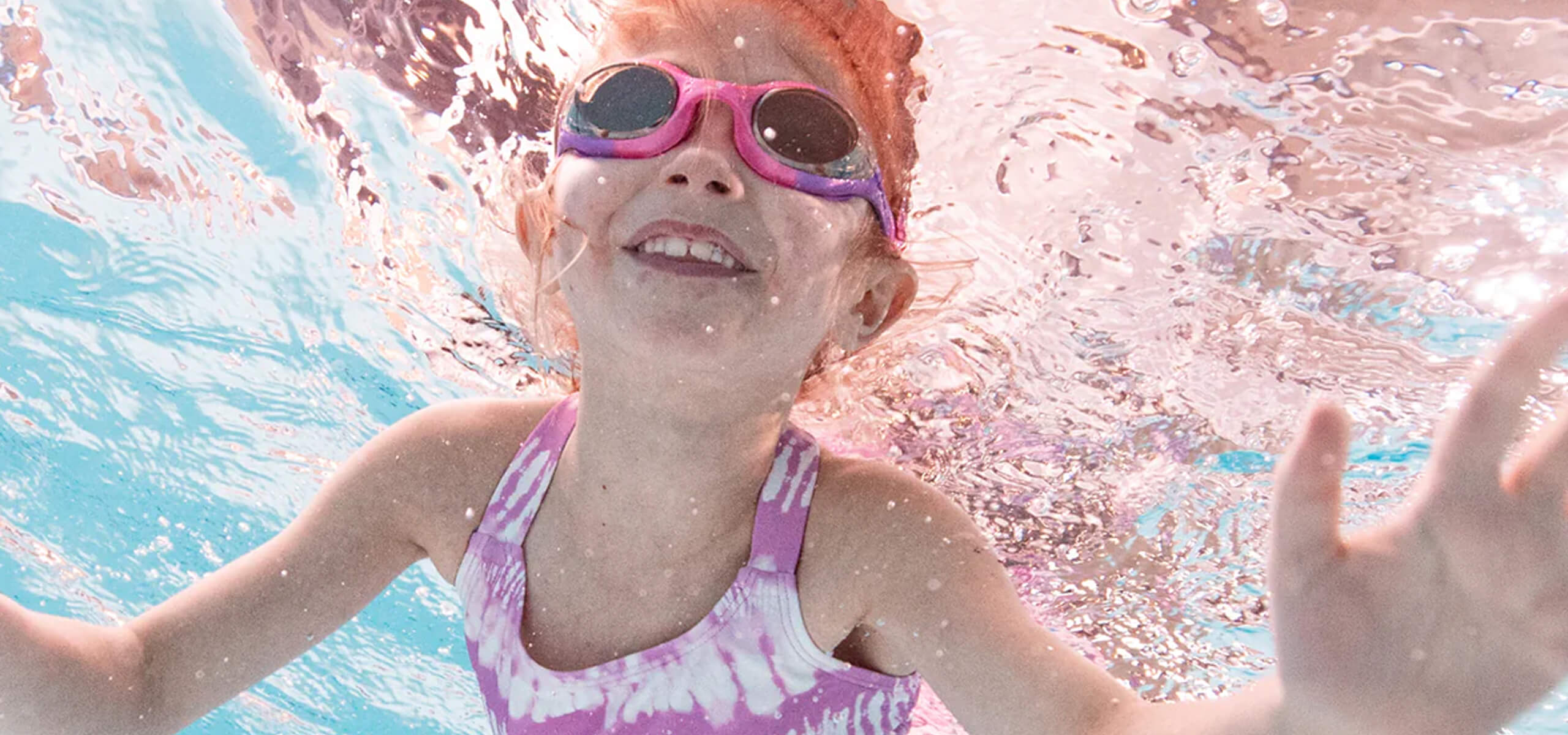 Photo of a child swimming underwater with Splash About pink and purple goggles, and a purple tie-dye swimsuit.